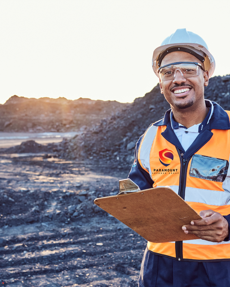 A smiling industrial worker wearing safety gear, including a hard hat, safety glasses, and a high-visibility vest, holding a clipboard at a mining or construction site with piles of excavated material in the background.