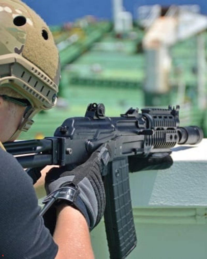 A tactical security operative wearing a helmet and gloves aims a rifle while positioned on a ship, indicating a maritime security operation. The background shows the deck of a vessel at sea.