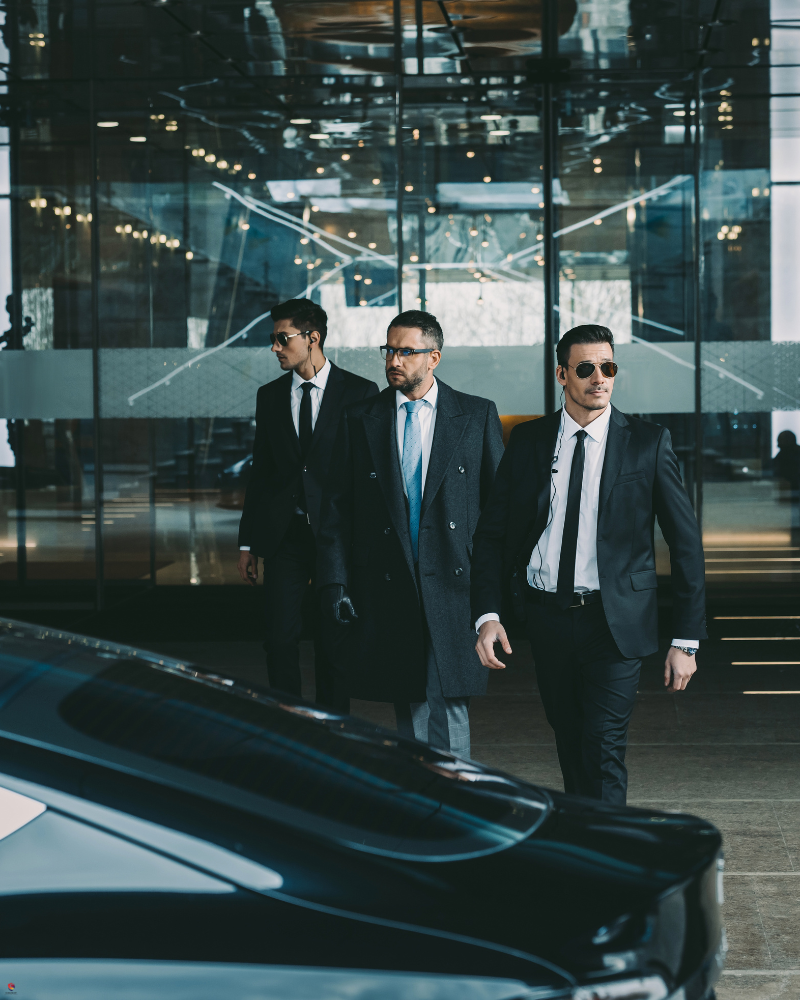 Three men in suits, walking purposefully in front of a glass building, with a sleek black car in the foreground.