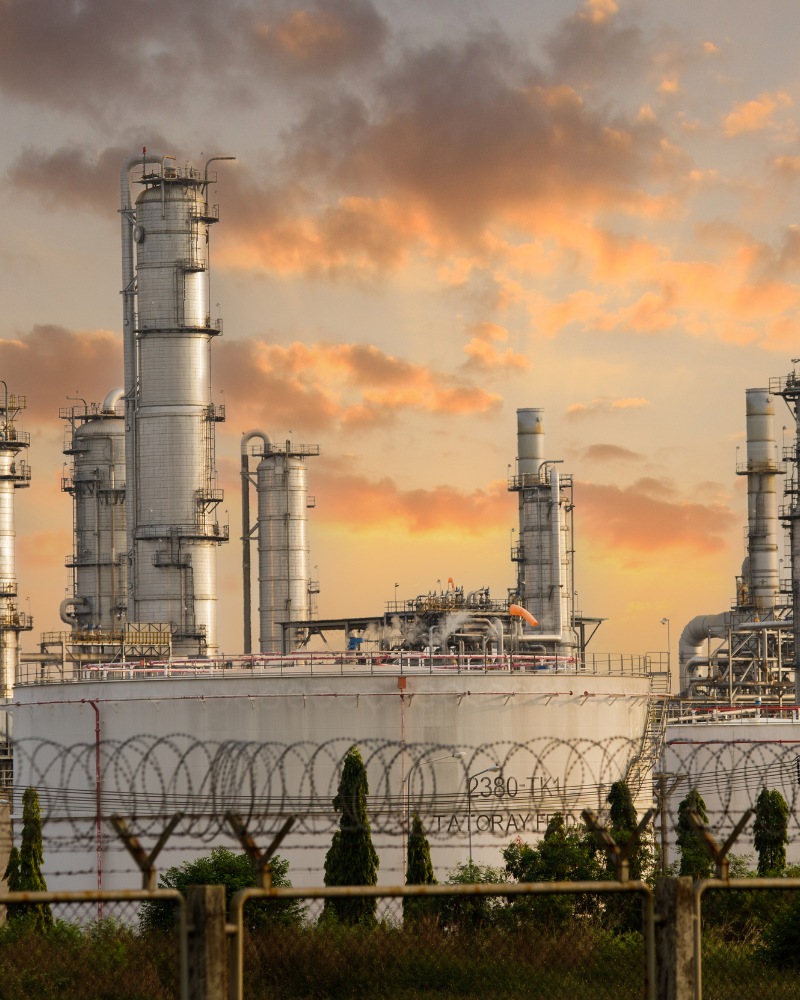 An industrial facility with tall cylindrical towers and pipelines, surrounded by a barbed wire fence, set against a sunset sky.