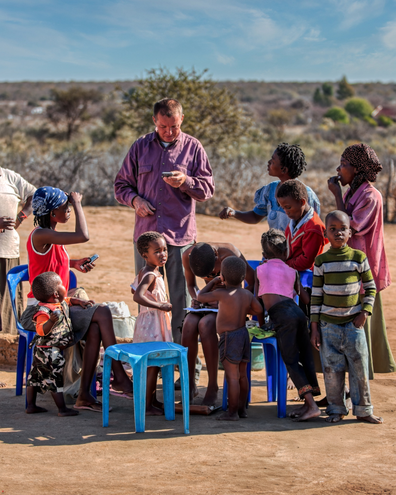 A group of children and adults gathered around a man distributing items in an outdoor rural setting. Some children are sitting on small blue plastic chairs while others stand nearby. The background features dry terrain with scattered trees and simple dwellings.